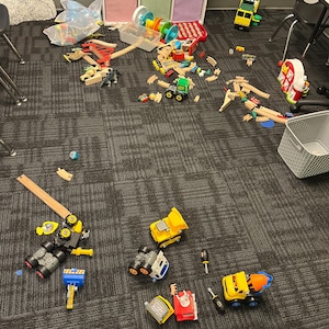 May include: A collection of children's toys scattered on a carpeted floor. The toys include wooden train tracks, toy trucks, and a variety of other colorful playthings. A storage unit with colorful cubbies is in the background.