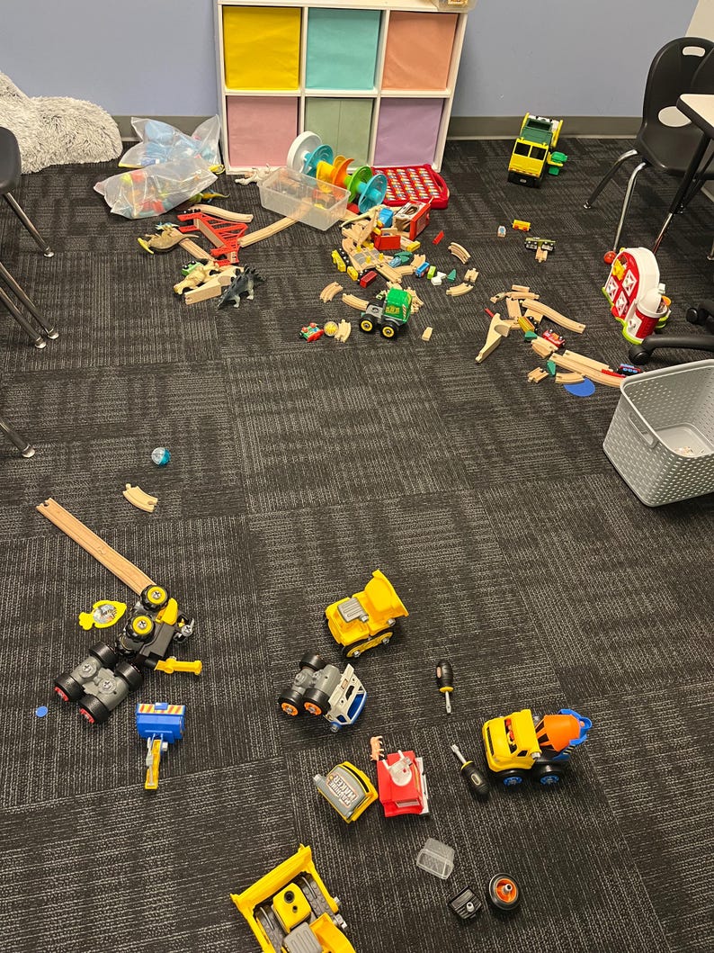May include: A collection of children's toys scattered on a carpeted floor. The toys include wooden train tracks, toy trucks, and a variety of other colorful playthings. A storage unit with colorful cubbies is in the background.