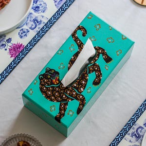 May include: Teal tissue box cover with a black and brown leopard print design. The leopard is depicted in a playful pose with its head turned to the side. The cover has a white tissue box insert.