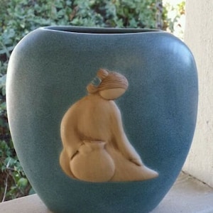 May include: A teal-colored ceramic vase with a textured finish. The vase features a raised relief of a seated figure holding a small pot. The figure is light beige, contrasting with the blue-green vase. A decorative piece for home decor.