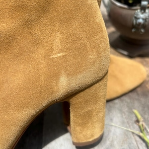 May include: Close-up of a tan suede boot. The boot features a block heel and a soft, textured surface. The color is a warm, earthy tone, suggesting a fall or winter style. The focus is on the texture and shape of the footwear.