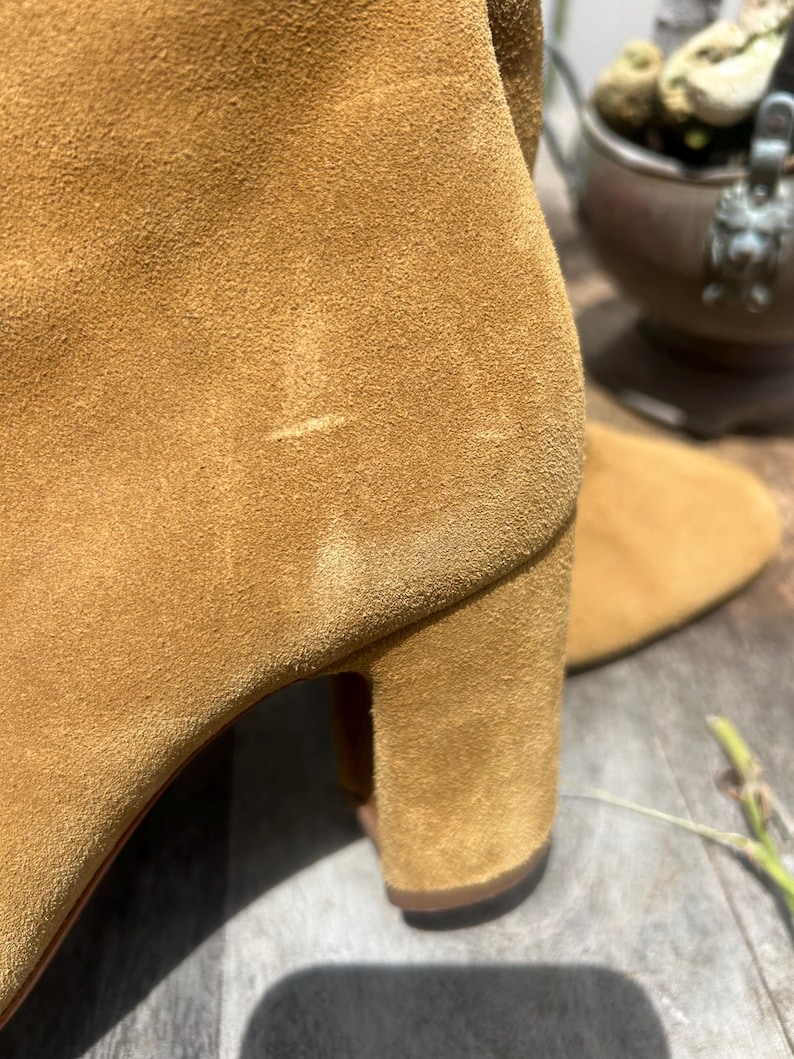 May include: Close-up of a tan suede boot. The boot features a block heel and a soft, textured surface. The color is a warm, earthy tone, suggesting a fall or winter style. The focus is on the texture and shape of the footwear.