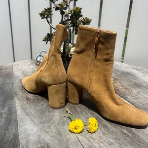 May include: A pair of tan suede ankle boots with block heels. One boot has a side zipper. The boots are set on a wooden surface with two yellow flowers and a small plant in the background. The boots have a classic, vintage style.