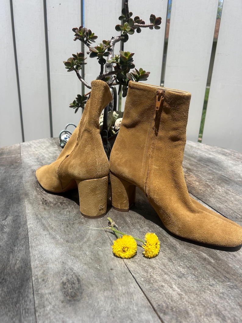 May include: A pair of tan suede ankle boots with block heels. One boot has a side zipper. The boots are set on a wooden surface with two yellow flowers and a small plant in the background. The boots have a classic, vintage style.