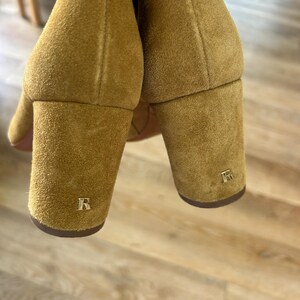 May include: Close-up of two mustard-colored suede boots with block heels. Each heel features a gold-colored letter "R". The boots are set against a light-colored wooden floor.