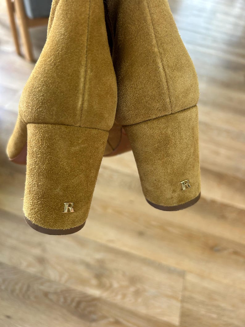 May include: Close-up of two mustard-colored suede boots with block heels. Each heel features a gold-colored letter "R". The boots are set against a light-colored wooden floor.