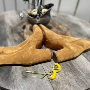 May include: A pair of tan suede ankle boots with a pointed toe and a small heel. The boots are laid out on a wooden surface. A small yellow flower and a decorative metal container are also present.