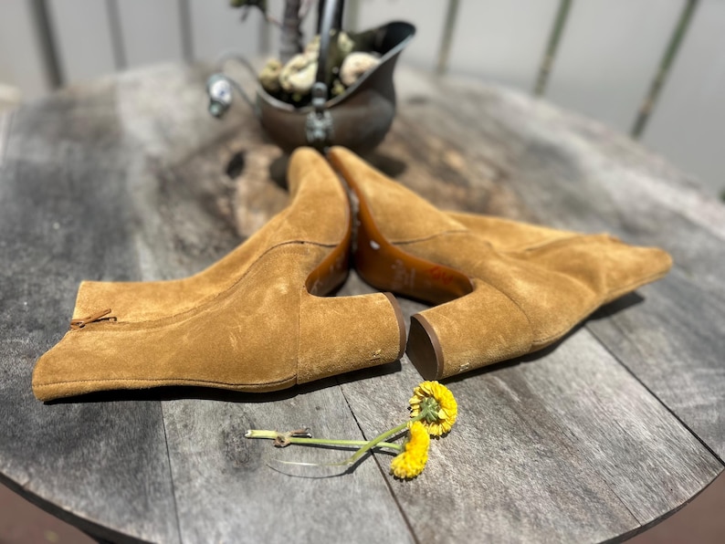 May include: A pair of tan suede ankle boots with a pointed toe and a small heel. The boots are laid out on a wooden surface. A small yellow flower and a decorative metal container are also present.