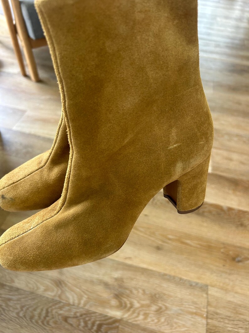 May include: Mustard-colored suede ankle boots with a rounded toe and a block heel. The boots have a mid-calf height and a simple, classic design. The heels measure approximately 3 inches.