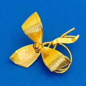 May include: A gold-toned brooch in the shape of a flower with three petals. The brooch has a textured surface and a small, round centre.