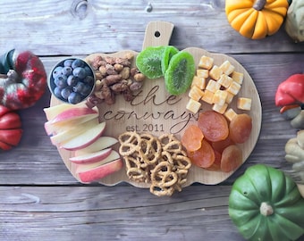 May include: A wooden pumpkin-shaped serving board with a variety of snacks, including dried fruit, pretzels, cheese, kiwi, apples, and nuts. The board is engraved with the text "the conways est. 2011".