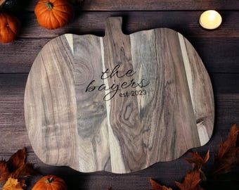 May include: A wooden pumpkin-shaped cutting board with the text "the bayers est. 2023" engraved on the surface. The board is surrounded by fall leaves and pumpkins.