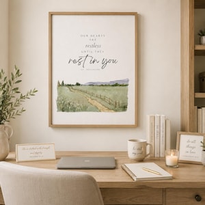 May include: A framed wall art print featuring a watercolor landscape of a dirt path through a green field with a wooden fence, accompanied by the text: Our hearts are restless until they rest in you, St. Augustine, in a light wood frame.