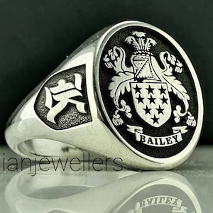 Może przedstawiać: Srebrny pierścionek z czarnym, owalnym herbem przedstawiającym herb ze tarczą, gwiazdami i tekstem "BAILEY". Pierścionek ma również tarczowaty wzór z boku z chińskim znakiem "天".