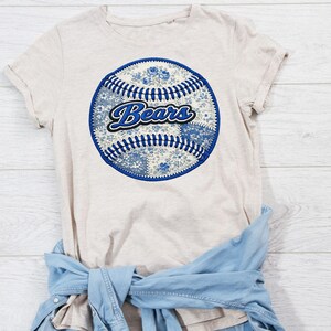 May include: Beige t-shirt featuring a baseball design with a blue floral pattern and the word "Bears" in a script font. The baseball has blue stitching. A light blue denim shirt is tied around the waist.