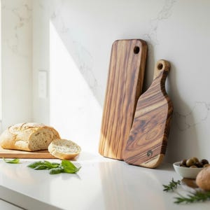May include: Two wooden chopping boards with a natural wood grain. One is rectangular with a hanging hole, the other has a handle. A loaf of bread and basil leaves are on the counter, suggesting a kitchen setting.