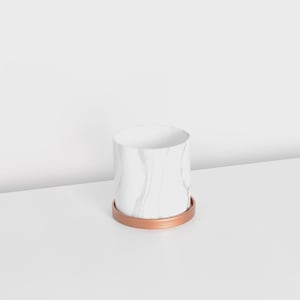 May include: A white marble-patterned ceramic planter with a copper-colored base. The cylindrical pot has a smooth, glossy finish and sits on a matching round saucer. The planter is designed for indoor use and adds a touch of modern elegance.
