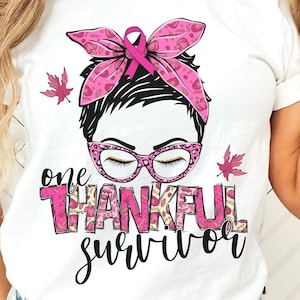 May include: White t-shirt with a pink and black graphic of a woman with a pink ribbon in her hair. The text "one thankful survivor" is printed on the shirt.