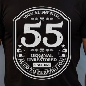 May include: Black t-shirt with a white graphic design. The graphic features a vintage-style badge with the number "55" in the center, surrounded by the text "100% Authentic Original & Unrestored Since 1970 Aged to Perfection".