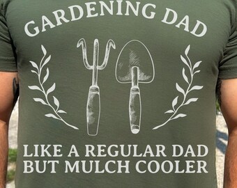 Op de afbeelding: Een groen T-shirt met de tekst "Gardening Dad Like a Regular Dad But Mulch Cooler" en een afbeelding van tuingereedschap. De tekst is wit en de gereedschappen zijn wit met een grijze omtrek. Het shirt is legergroen.
