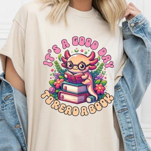 May include: Beige t-shirt with a colorful graphic of an axolotl wearing glasses and reading a book. The text "It's a good day to read a book" is in a circular design around the axolotl. The design includes flowers and leaves.