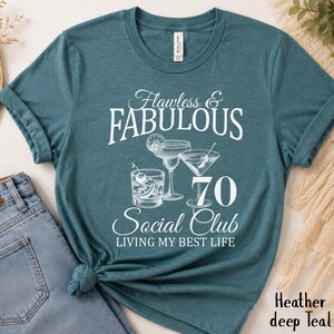 May include: Teal t-shirt with the text "Flawless & Fabulous 70 Social Club Living My Best Life". The shirt features illustrations of cocktails. The shirt is made of a soft material.