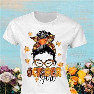 May include: White t-shirt with a graphic of a woman with a messy bun, wearing glasses and a headband with fall leaves and a pumpkin. The text "October Girl" is written in fall colors below the woman's face.