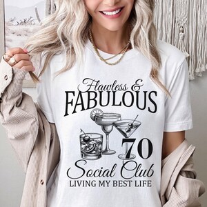 May include: White t-shirt with the text "Flawless & Fabulous 70 Social Club Living My Best Life." The shirt features illustrations of cocktails. The design is in black.