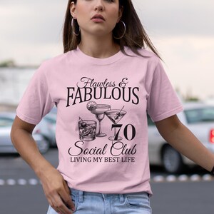 May include: A light pink t-shirt with the text "Flawless & Fabulous" above a graphic of cocktails and the number 70. Below the graphic, the text reads "Social Club, Living My Best Life."