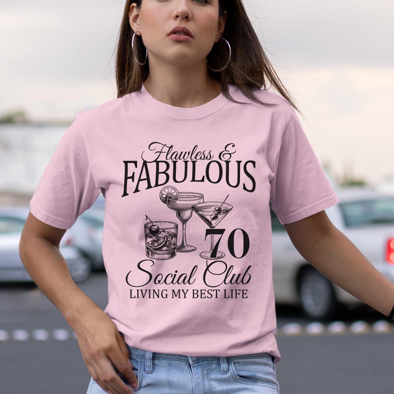 May include: A light pink t-shirt with the text "Flawless & Fabulous" above a graphic of cocktails and the number 70. Below the graphic, the text reads "Social Club, Living My Best Life."