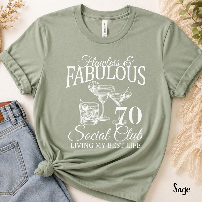 May include: Sage green t-shirt with the text "Flawless & Fabulous 70 Social Club Living My Best Life." The design includes illustrations of cocktails. The shirt is made of a soft material.