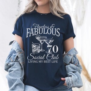 May include: Navy blue t-shirt with the text "Flawless & Fabulous 70 Social Club Living My Best Life". The shirt features cocktail illustrations in white. A denim jacket is draped over the shoulders.