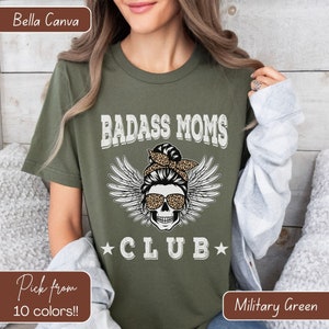 May include: A green t-shirt with the text "BADASS MOMS CLUB" and a skull with wings and a messy bun wearing sunglasses. The skull is wearing a leopard print bandana.