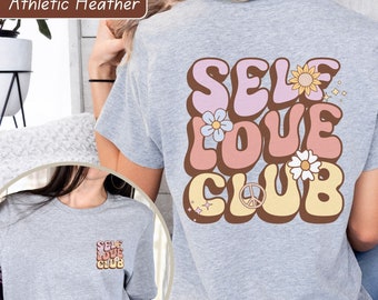 Op de afbeelding: Athletic heather grijs t-shirt met de woorden "SELF LOVE CLUB" in een retro lettertype op de rug en een kleinere versie op de voorkant. Het ontwerp bevat bloemen en een vredesteken in roze, bruin en geel.