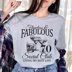 May include: A gray t-shirt with the words "Flawless & Fabulous" above the number "70" and the words "Social Club" and "Living My Best Life". The shirt features illustrations of cocktails.