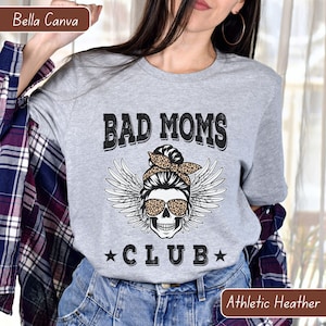 Puede incluir: Camiseta gris con el texto "Bad Moms Club" en negro. Un cr&aacute;neo con alas de &aacute;ngel y un pa&ntilde;uelo de estampado de leopardo se encuentra en el centro del texto.