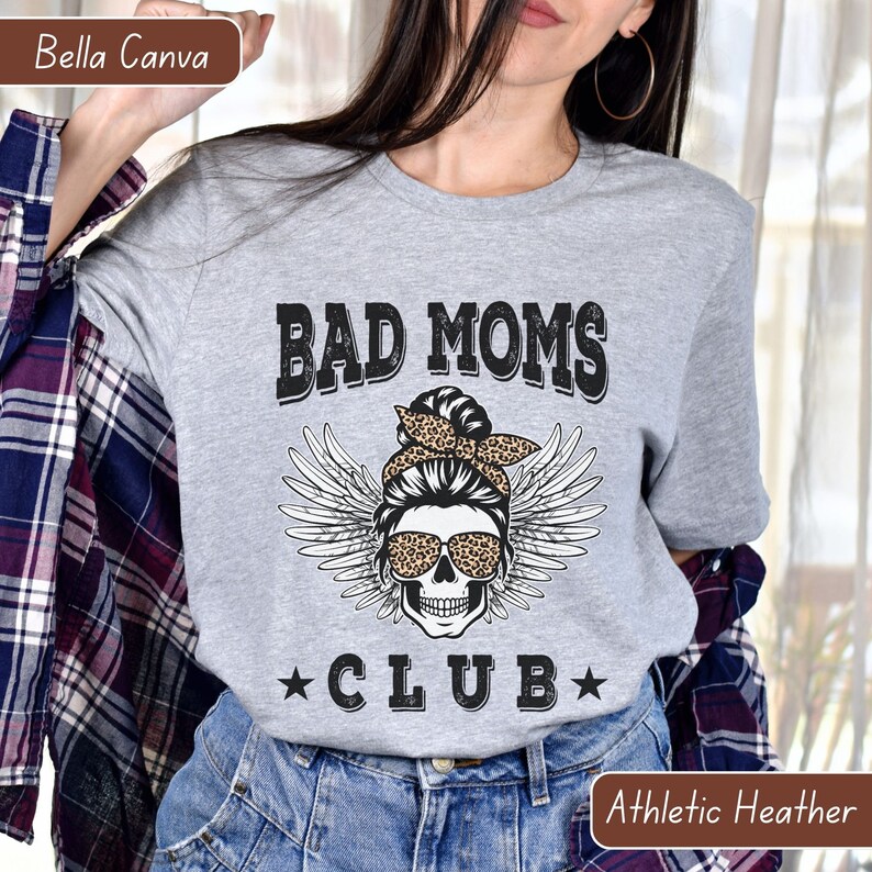 Puede incluir: Camiseta gris con el texto "Bad Moms Club" en negro. Un cr&aacute;neo con alas de &aacute;ngel y un pa&ntilde;uelo de estampado de leopardo se encuentra en el centro del texto.