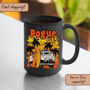 May include: A black ceramic coffee mug with a white van and surfboards on the side. The van has the text "Pogue life" in orange and yellow letters. The mug has a sunset background with palm trees. The mug is 11 oz or 15 oz.