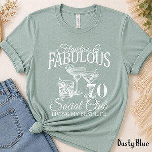 May include: Dusty blue t-shirt with the text "Flawless & Fabulous 70 Social Club Living My Best Life." The graphic includes illustrations of cocktails. The shirt is made of a soft material.