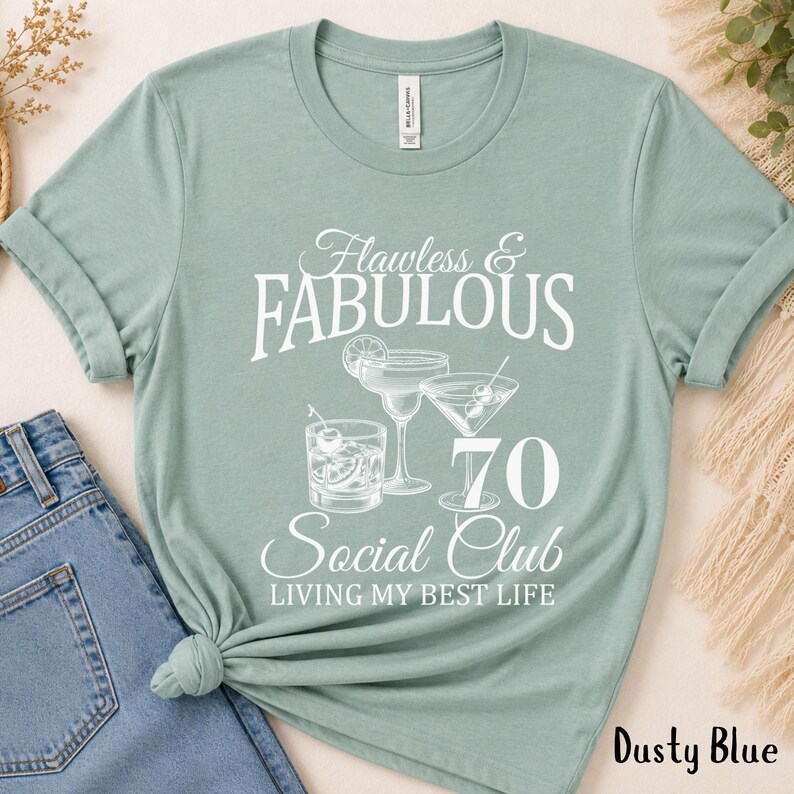 May include: Dusty blue t-shirt with the text "Flawless & Fabulous 70 Social Club Living My Best Life." The graphic includes illustrations of cocktails. The shirt is made of a soft material.