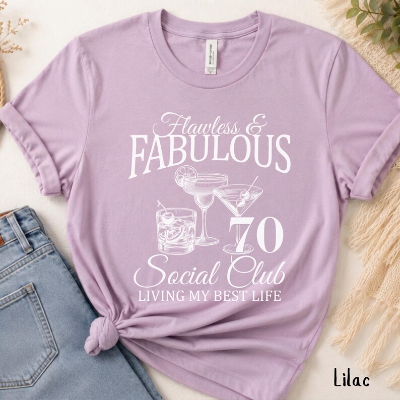 May include: Lilac-colored t-shirt with the text "Flawless & Fabulous 70 Social Club Living My Best Life." The design includes illustrations of cocktails. The shirt is paired with blue jeans.