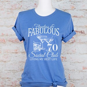 May include: A blue t-shirt with rolled-up sleeves. The shirt features the text "Flawless & Fabulous 70 Social Club" with illustrations of cocktails. The phrase "Living My Best Life" is also printed on the shirt.