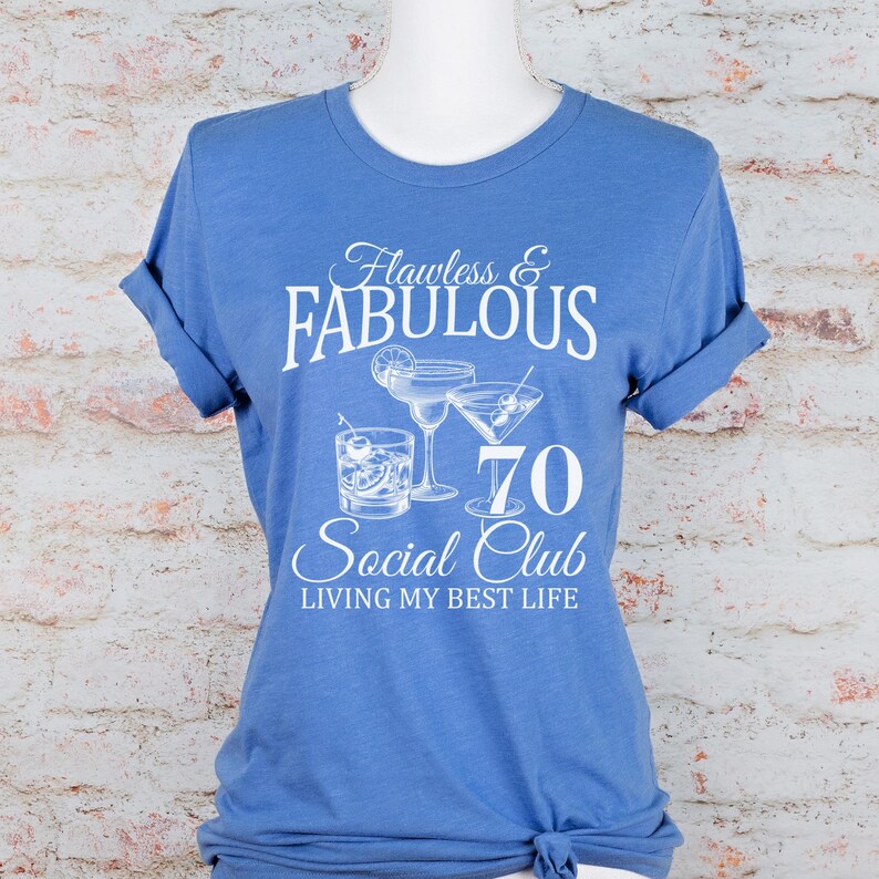 May include: A blue t-shirt with rolled-up sleeves. The shirt features the text "Flawless & Fabulous 70 Social Club" with illustrations of cocktails. The phrase "Living My Best Life" is also printed on the shirt.