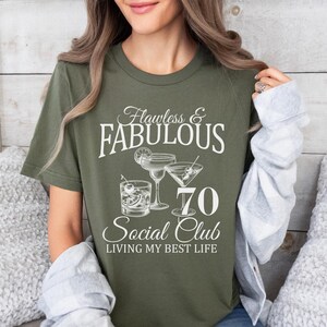 May include: Olive green t-shirt with the text "Flawless & Fabulous 70 Social Club Living My Best Life." The graphic includes illustrations of cocktails. A woman is wearing the shirt.