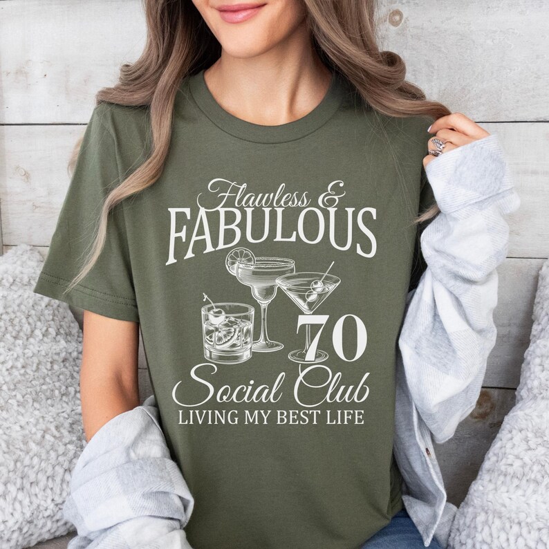 May include: Olive green t-shirt with the text "Flawless & Fabulous 70 Social Club Living My Best Life." The graphic includes illustrations of cocktails. A woman is wearing the shirt.