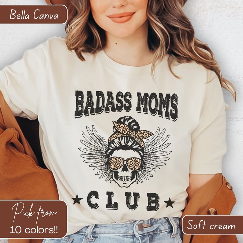 Puede incluir: Una camiseta blanca con el texto "Badass Moms Club" en letras negras. El dise&ntilde;o presenta una calavera con gafas de sol y un mo&ntilde;o desordenado con un pa&ntilde;uelo de estampado de leopardo. El texto "Pick from 10 colors!" est&aacute; en la esquina inferior izquierda y "Soft cream" est&aacute; en la esquina inferior derecha.