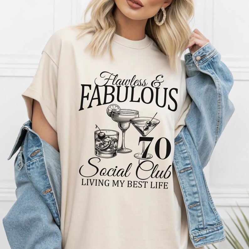 May include: A cream-colored t-shirt with the text "Flawless & Fabulous" above the words "70 Social Club" and "Living My Best Life." The shirt features illustrations of cocktails.