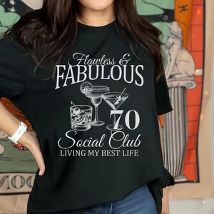 May include: Black t-shirt with the words "Flawless & Fabulous" above the text "70 Social Club" and the phrase "Living My Best Life." The shirt features illustrations of cocktails.