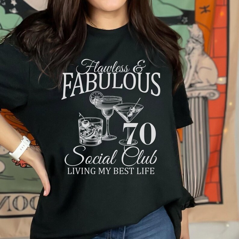 May include: Black t-shirt with the words "Flawless & Fabulous" above the text "70 Social Club" and the phrase "Living My Best Life." The shirt features illustrations of cocktails.