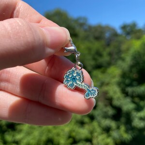 May include: A silver-toned metal charm shaped like a neuron cell, featuring light blue and white enamel detailing. The charm includes a small lobster claw clasp at the top for attachment, with a clear view of the dendrites and axon structure.